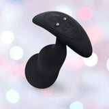 The B-Vibe Vibrating Snug Plug RC: Silicone P-Spot Anal Vibrator - Large by B-Vibe, featuring a curved black silicone design with control buttons and metal contact points, is set against a soft pastel background with blurred lights.