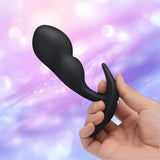 A hand holds the B-Vibe Vibrating P-Spot Snug Plug RC Large by B-Vibe, featuring a black, curved design, set against a softly blurred pastel background in pink, purple, and blue tones.