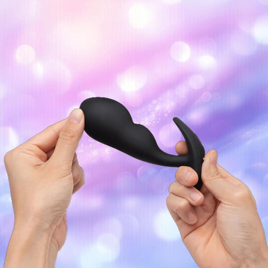 Two hands hold and stretch a black B-Vibe Vibrating P-Spot Snug Plug RC Large by B-Vibe against a blurred, pastel purple and blue background with bokeh lighting effects. 1080
