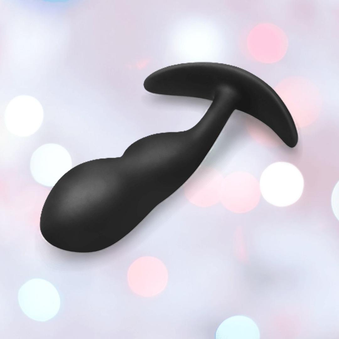 The B-Vibe Vibrating Snug Plug RC: Silicone P-Spot Anal Vibrator - Large by B-Vibe, in black silicone and whale-tail shaped, is displayed against a pastel bokeh background.