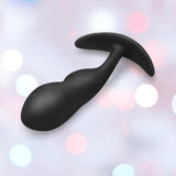 The B-Vibe Vibrating Snug Plug RC: Silicone P-Spot Anal Vibrator - Large by B-Vibe, in black silicone and whale-tail shaped, is displayed against a pastel bokeh background.