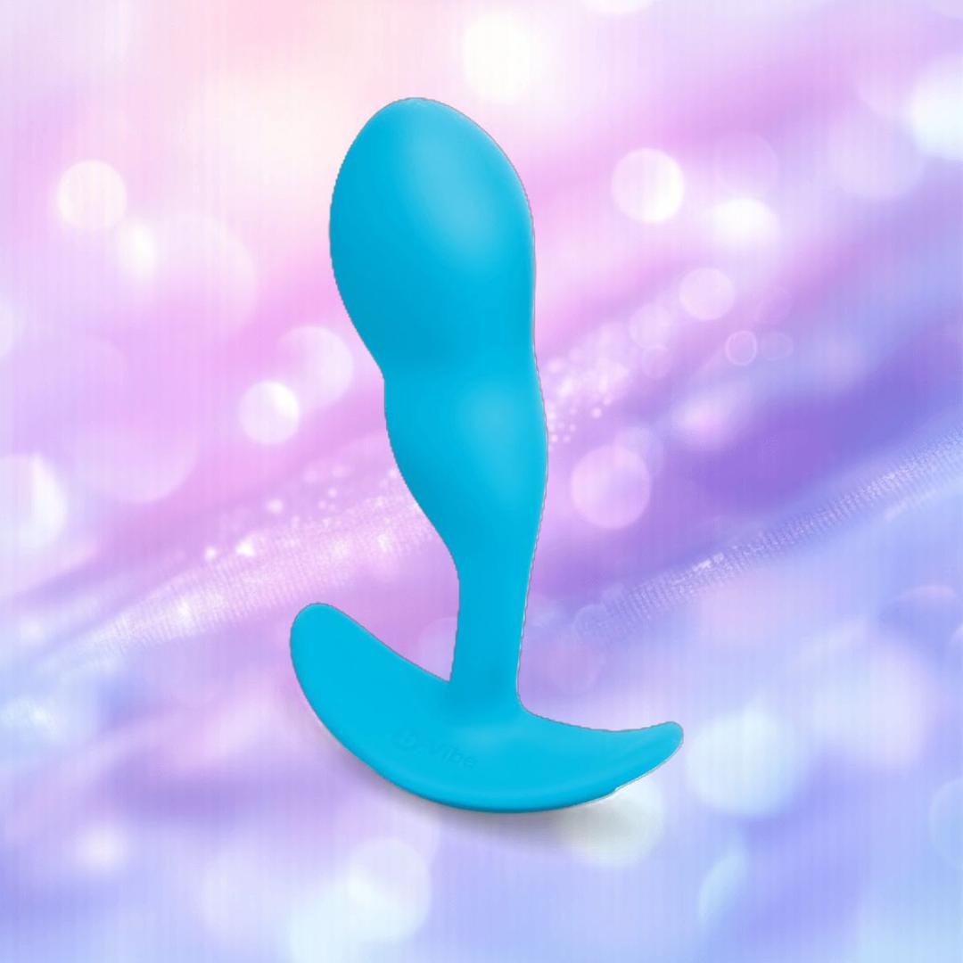 The B-Vibe Vibrating P-Spot Snug Plug RC Small, a blue silicone prostate massager with an ergonomic curve, stands upright on a dreamy pastel purple and pink background with glowing bokeh, designed by B-Vibe for effortless stimulation.