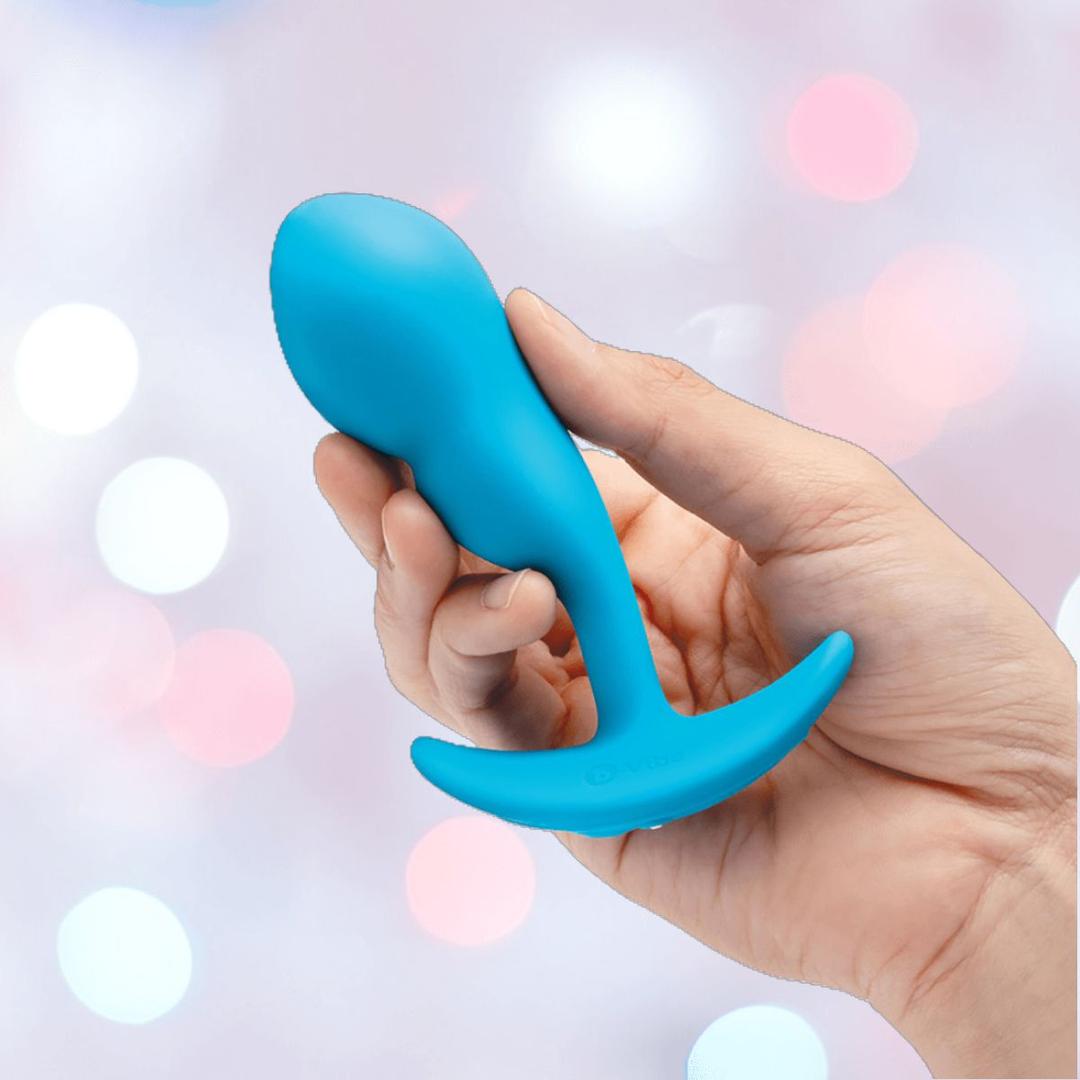 A hand holds the B-Vibe Vibrating Snug Plug RC: Silicone P-Spot Anal Vibrator (Small) by B-Vibe, featuring a blue curved base, against a soft pastel bokeh background.