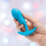 A hand holds the B-Vibe Vibrating Snug Plug RC: Silicone P-Spot Anal Vibrator (Small) by B-Vibe, featuring a blue curved base, against a soft pastel bokeh background.