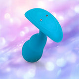 The B-Vibe Vibrating P-Spot Snug Plug RC Small by B-Vibe, a blue silicone vibrating butt plug with a flat oval base and two metallic contact points for prostate stimulation, is shown against a soft pastel pink and purple bokeh background.