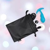 A black drawstring bag sits open, revealing the B-Vibe Vibrating Snug Plug RC: Silicone P-Spot Anal Vibrator - Small by B-Vibe, plus its remote control and USB charging cable, set against a soft pastel background.