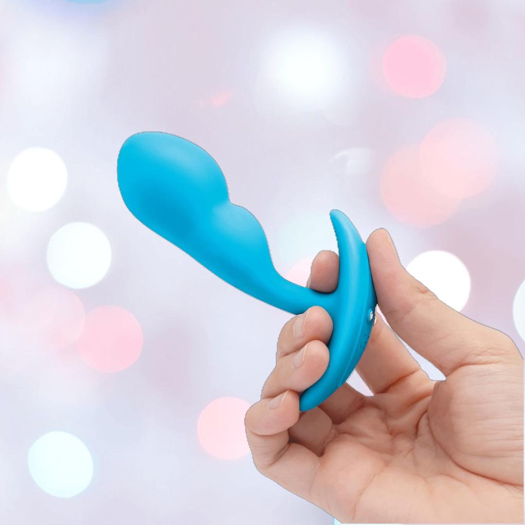 A hand holds the B-Vibe Vibrating Snug Plug RC (Small), a blue silicone P-spot anal vibrator with a curved, ergonomic design, against a blurred backdrop of soft pink and white bokeh lights.