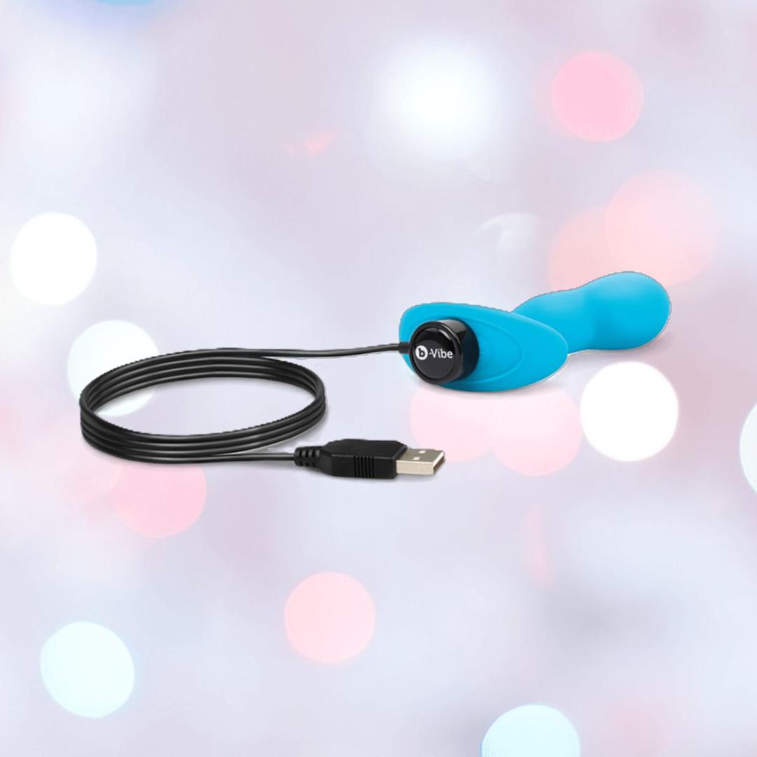 The B-Vibe Vibrating Snug Plug RC: Silicone P-Spot Anal Vibrator - Small by B-Vibe in blue, with a USB charging cable attached, is shown against a softly blurred backdrop of pink, white, and blue bokeh lights.