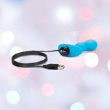 The B-Vibe Vibrating Snug Plug RC: Silicone P-Spot Anal Vibrator - Small by B-Vibe in blue, with a USB charging cable attached, is shown against a softly blurred backdrop of pink, white, and blue bokeh lights.