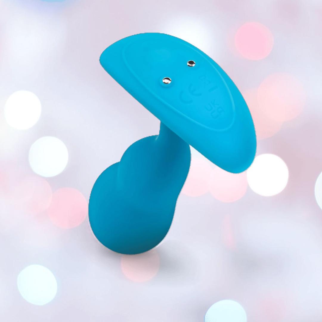 The B-Vibe Vibrating Snug Plug RC: Silicone P-Spot Anal Vibrator - Small is a blue B-Vibe silicone plug with a rounded base, flat oval top, and two metal contacts, displayed against a soft pastel bokeh background.
