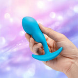 A hand holds the B-Vibe Vibrating P-Spot Snug Plug RC Small by B-Vibe against a soft, blurred background with purple and pink hues and light bokeh effects.