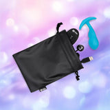 A black drawstring pouch reveals the B-Vibe Vibrating P-Spot Snug Plug RC Small, its USB charging cable, and a black remote control, all by B-Vibe, on a blurred purple and pink background.
