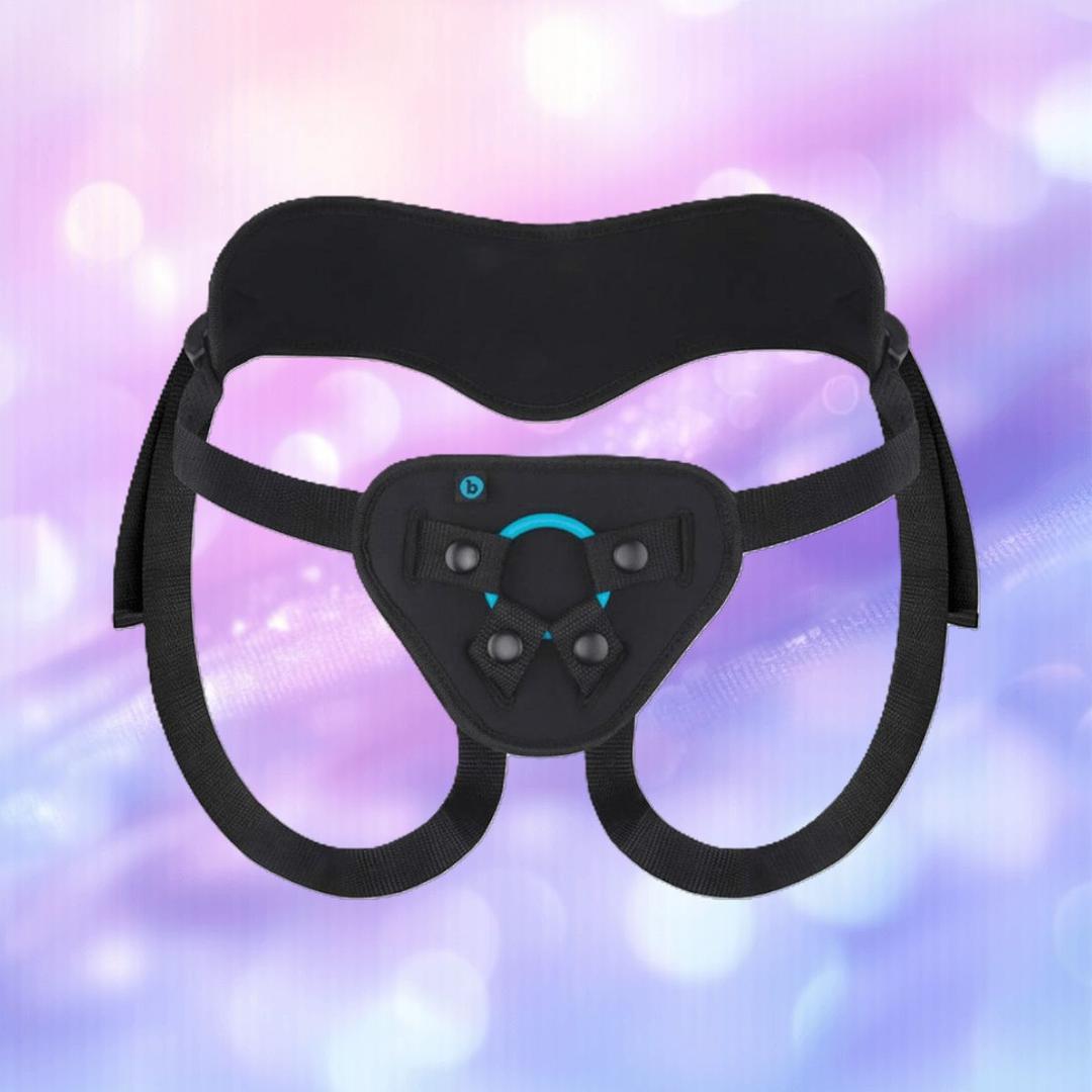 The B-Vibe Every Body Strap-On Harness Set with 6" Silicone Dildo features adjustable black straps and a padded front panel, shown against a soft, pastel pink, purple, and blue blurred background.