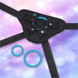 The B-Vibe Every Body Strap-On Harness Set with 6" Silicone Dildo by B-Vibe features a black adjustable harness with snap fasteners and three detachable blue silicone rings, ideal for pegging, shown on a pastel purple and blue background.