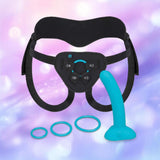 The B-Vibe Every Body Strap-On Harness Set with 6" Silicone Dildo by B-Vibe features a black adjustable harness, blue silicone dildo, and three blue rings, displayed on a soft purple and pink background—ideal for pegging adventures.