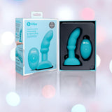 The B-Vibe Remote Control Rimming P-Spot Plug by B-Vibe and its remote are displayed in packaging beside the product box, featuring rotating beads for enhanced prostate stimulation, all set against a softly lit, blurred background.