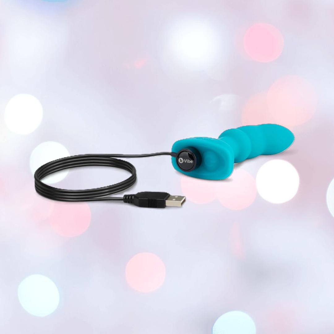 The B-Vibe Remote Control Rimming P-Spot Plug by B-Vibe is a blue, silicone USB-rechargeable prostate vibrator with flexible rotating beads and an attached charging cable, shown on a pastel background with bokeh light effects.