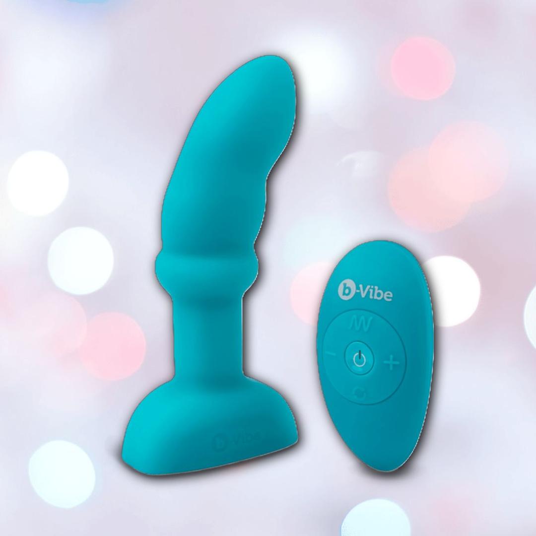 The B-Vibe Remote Control Rimming P-Spot Plug, a blue silicone prostate massager with a curved tip and flared base, sits next to its matching remote. Soft pink and white bokeh lights illuminate the background.