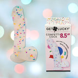 A novelty silicone dildo filled with colorful confetti, designed to resemble male genitalia, is positioned next to its packaging. The box is labeled "Get Lucky Confetti Cock 8.5''" and features a vibrant, playful design from the Get Lucky brand. The softly blurred background with pastel lights enhances the whimsical ambiance.