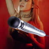 A person stands with a relaxed expression and closed eyes, wearing a choker necklace against a red backdrop. A large silver and black microphone is prominently featured in the foreground, reminiscent of the Doxy Die Cast 3 Wand Massager in Brushed Metal by Doxy, known for its deep, rumbly vibration.