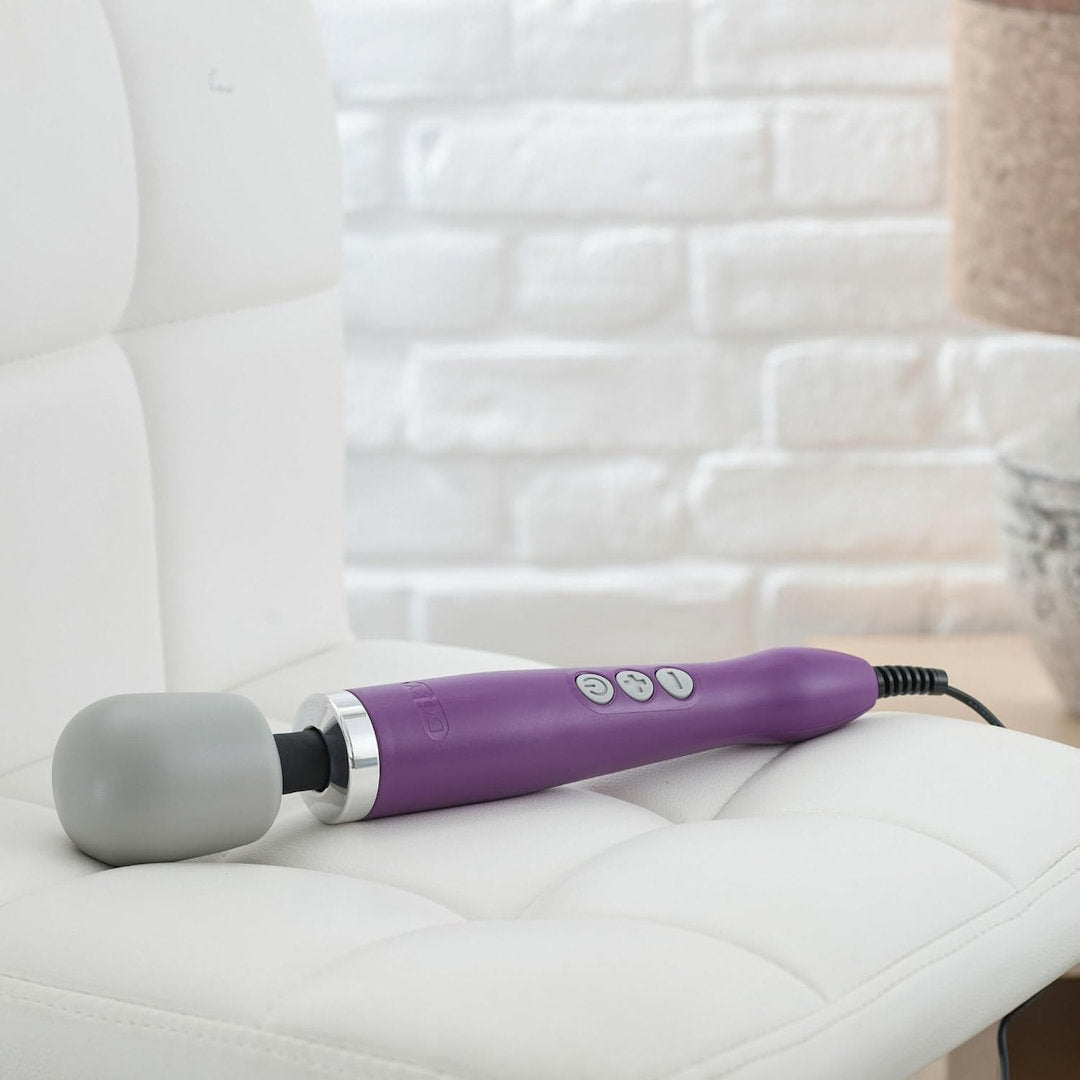 A Doxy Original Wand Massager - Purple, complete with buttons and a gray rounded head, rests on a white cushioned chair. The background showcases a white brick wall and a partial view of a table adorned with a decorative item. This Doxy handheld device delivers intense vibrations and an escalating pulse mode for ultimate relaxation.