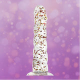 The Icon Brands Glittergasm Dong - Red and Orange Swirl 8" stands upright against a purple background with bokeh light effects, showcasing its clear, textured design containing small flower petals.