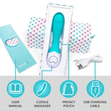 The OhMiBod Lovelife Cuddle G-Spot Vibrator in Turquoise is displayed inside an open box, showcasing its accessories: a user manual, a privacy pouch, and a USB charging cable. The packaging is white adorned with colorful heart patterns, and there is a heart-shaped detail on the base of the massager. Perfect for personal pleasure with targeted sensations.