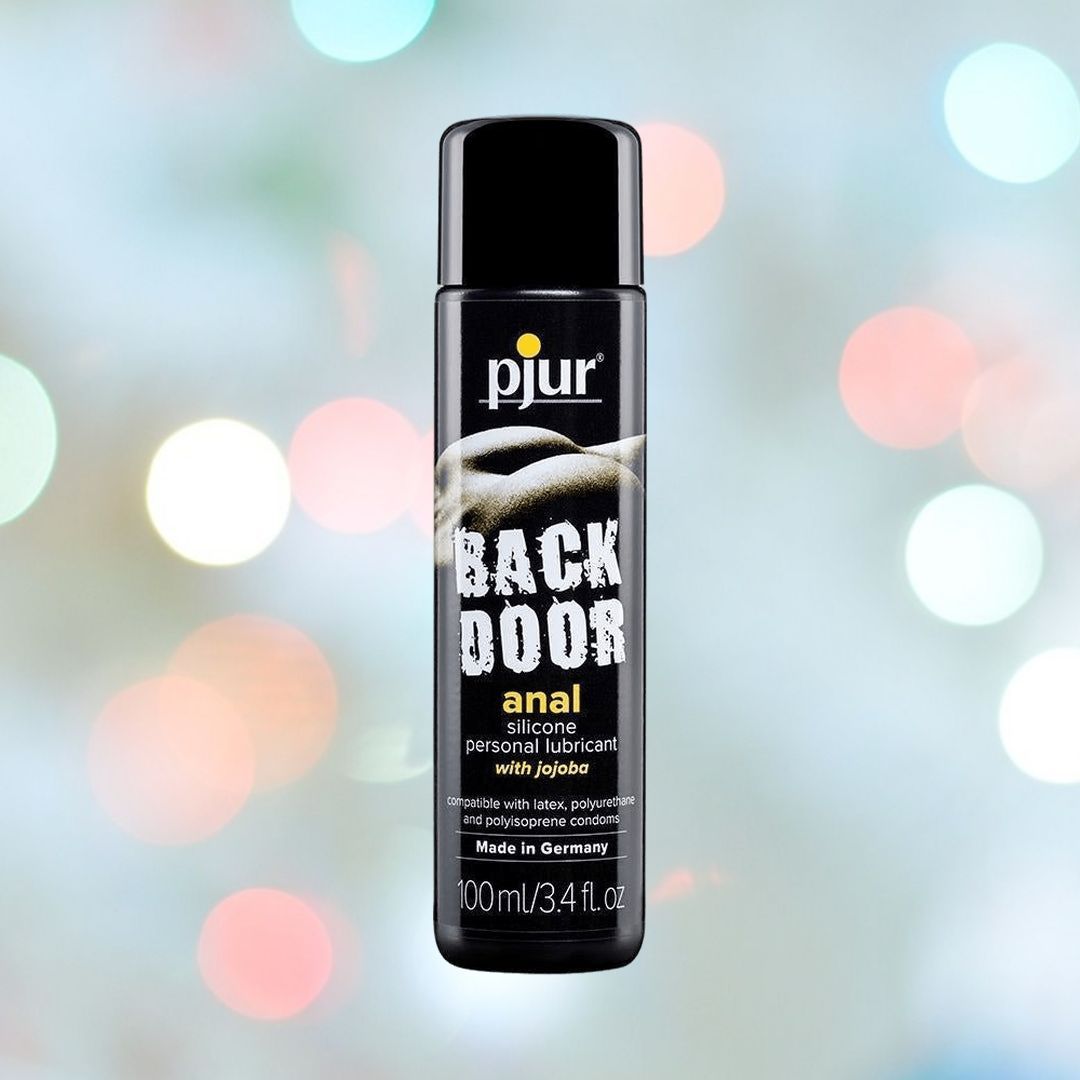 A bottle of Pjur Backdoor Relaxing Silicone Anal Glide premium anal lubricant with jojoba is shown. The label is black with the product name in bold white letters and other information in smaller text. The bottle size is 100 ml (3.4 fl. oz). The background is abstract and colorful.