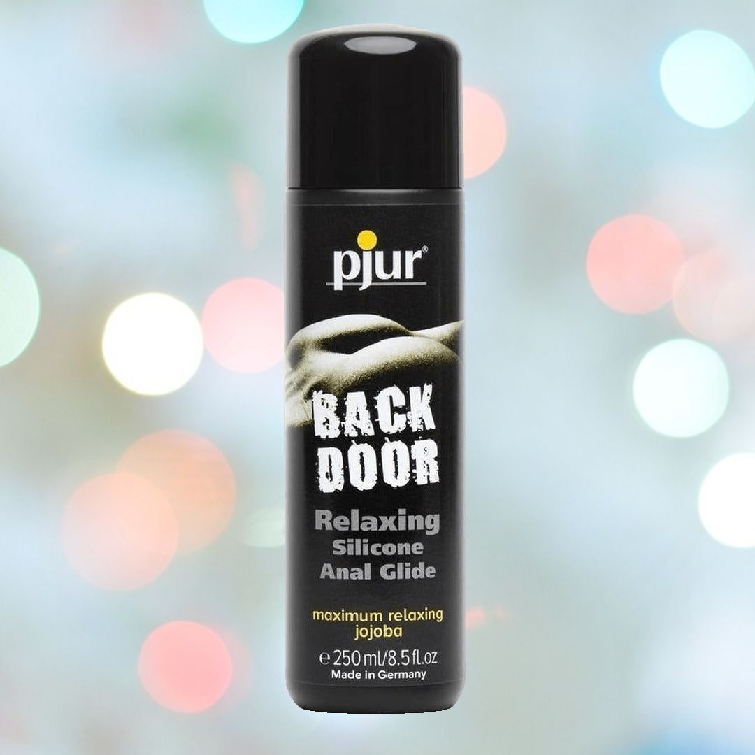 A black bottle of Pjur Backdoor Relaxing Silicone Anal Glide, a premium silicone anal lubricant from Pjur, is displayed against a blurred background featuring multicolored circular lights. The text on the bottle highlights it as a relaxing silicone-based product with jojoba. The bottle has a capacity of 250 ml (8.5 fl. oz).