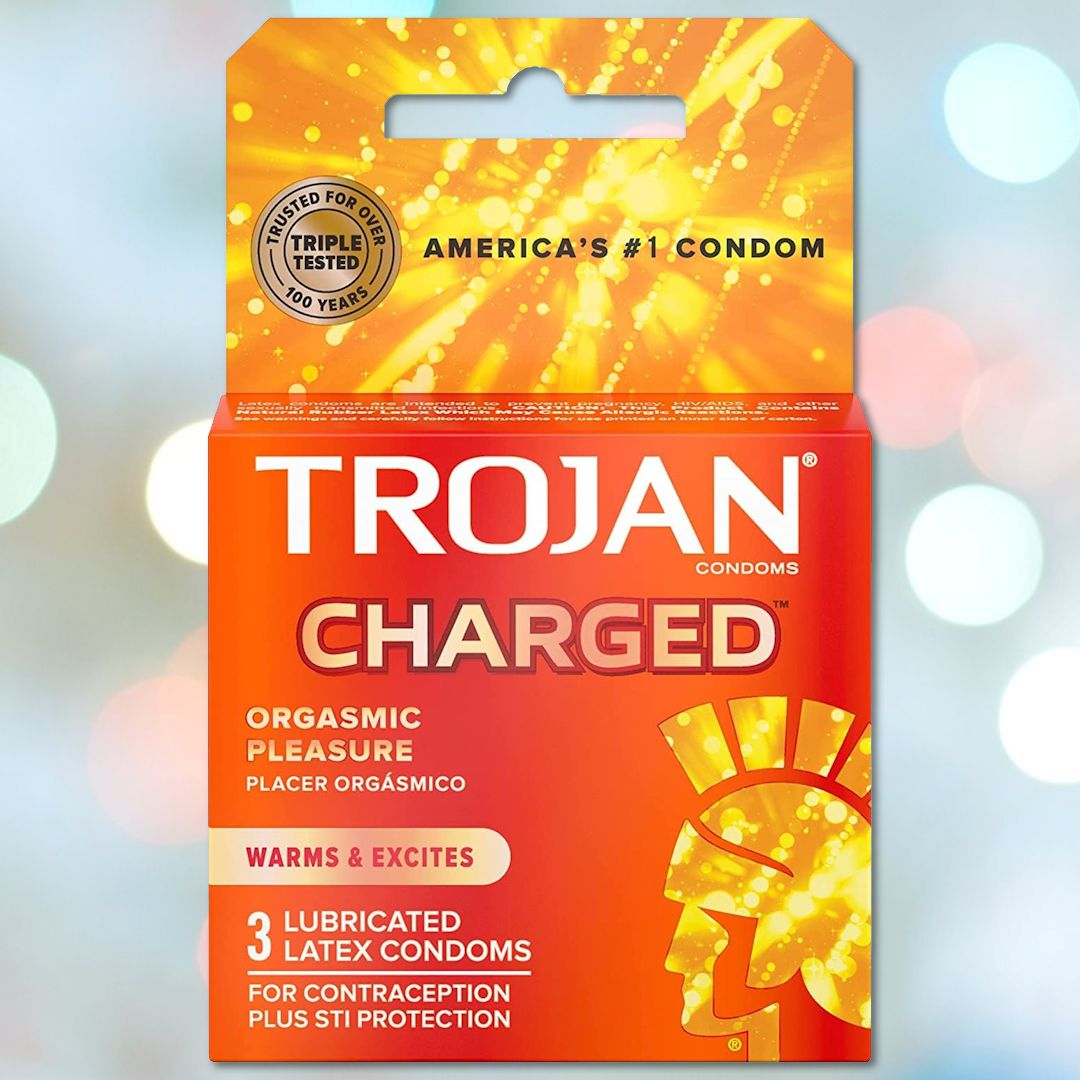 A package of Trojan Charged Deep-Ribbed Condoms is showcased against a blurred blue and white background. The orange and red packaging, featuring an orgasmic design, emphasizes features such as "orgasmic pleasure" and "warms & excites." This comfort fit pack includes three lubricated latex condoms.