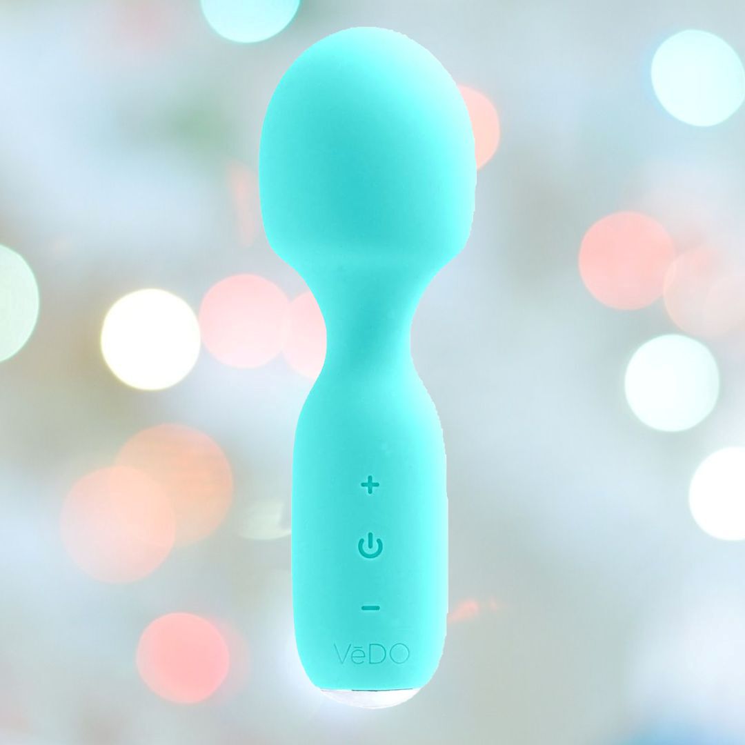 The VeDO WINI Mini Vibrator Wand in turquoise, with a rounded head and a handle adorned with control buttons, is displayed against a softly blurred background featuring circular light bokeh in shades of white, pink, and blue. This rechargeable mini wand by VeDO offers 10 vibration modes for personalized relaxation.