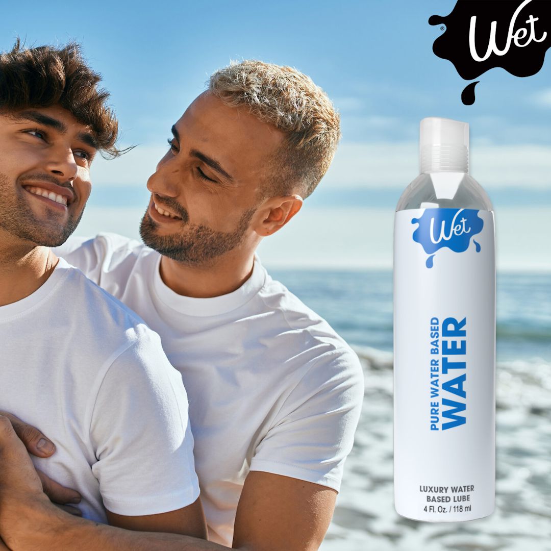 Two people share a joyful moment outdoors, as one hugs the other from behind, radiating sheer delight. In the foreground stands a bottle of Wet Water-Based Lubricant. The tranquil sky and water create a serene backdrop.