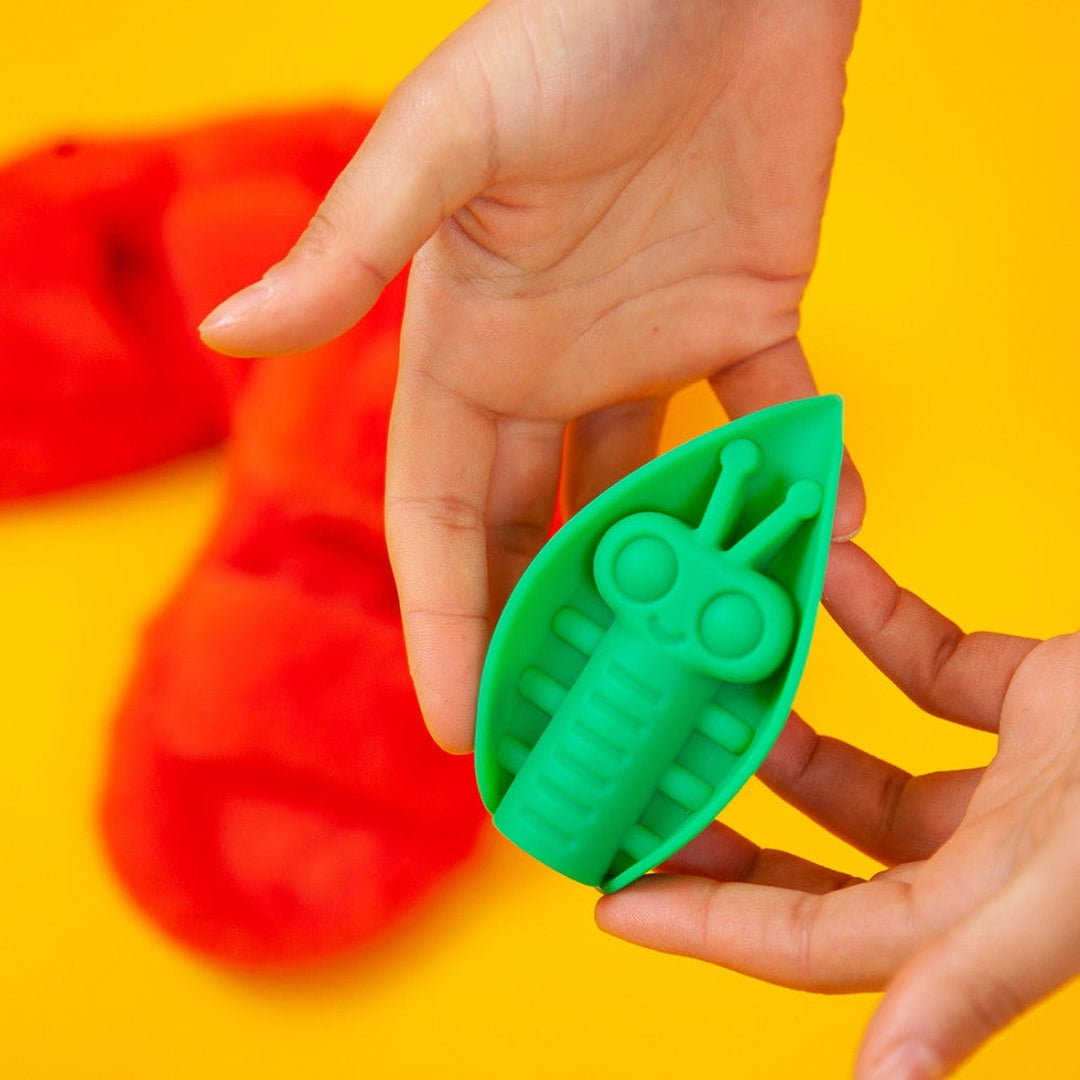 Hands holding an Emerald Cute Little Fuckers Zeep against a vibrant yellow background, with pulsating red dough in the backdrop.