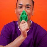 A person wearing a purple shirt holds a green Cute Little Fuckers Zeep - Emerald toy bug in front of their face against an orange background, as if peering into the vibrant mysteries of a jungle world.