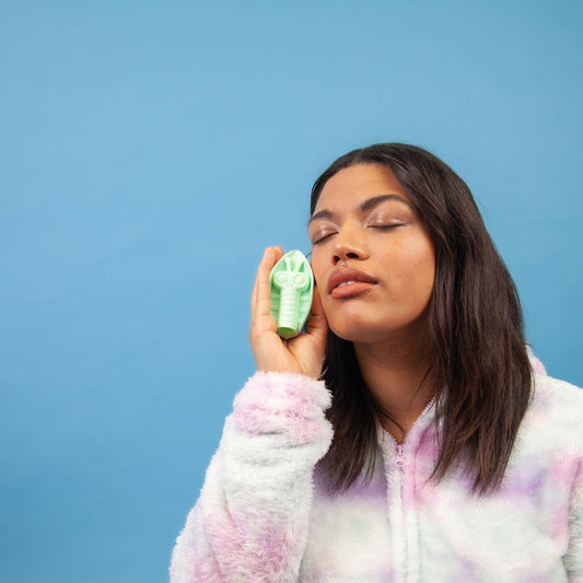 A woman with long dark hair, wearing a colorful tie-dye hoodie, holds the Cute Little Fuckers Zeep - Mint massage tool to her sensitive areas on her cheek, eyes closed, against a blue background. 1080