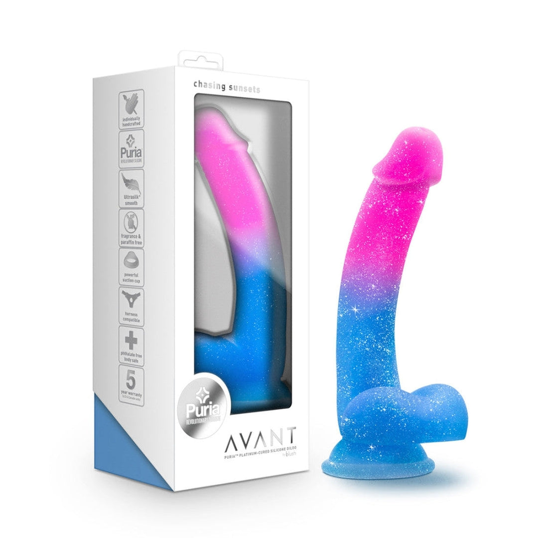 Image of the Avant Chasing Sunsets Dildo - Mermaid, featuring a gradient color scheme transitioning from pink to blue and covered in a glitter-like texture. It stands upright beside its packaging, which highlights features such as "chasing sunsets," a suction cup base, Puria platinum-cured silicone, and a five-year warranty.