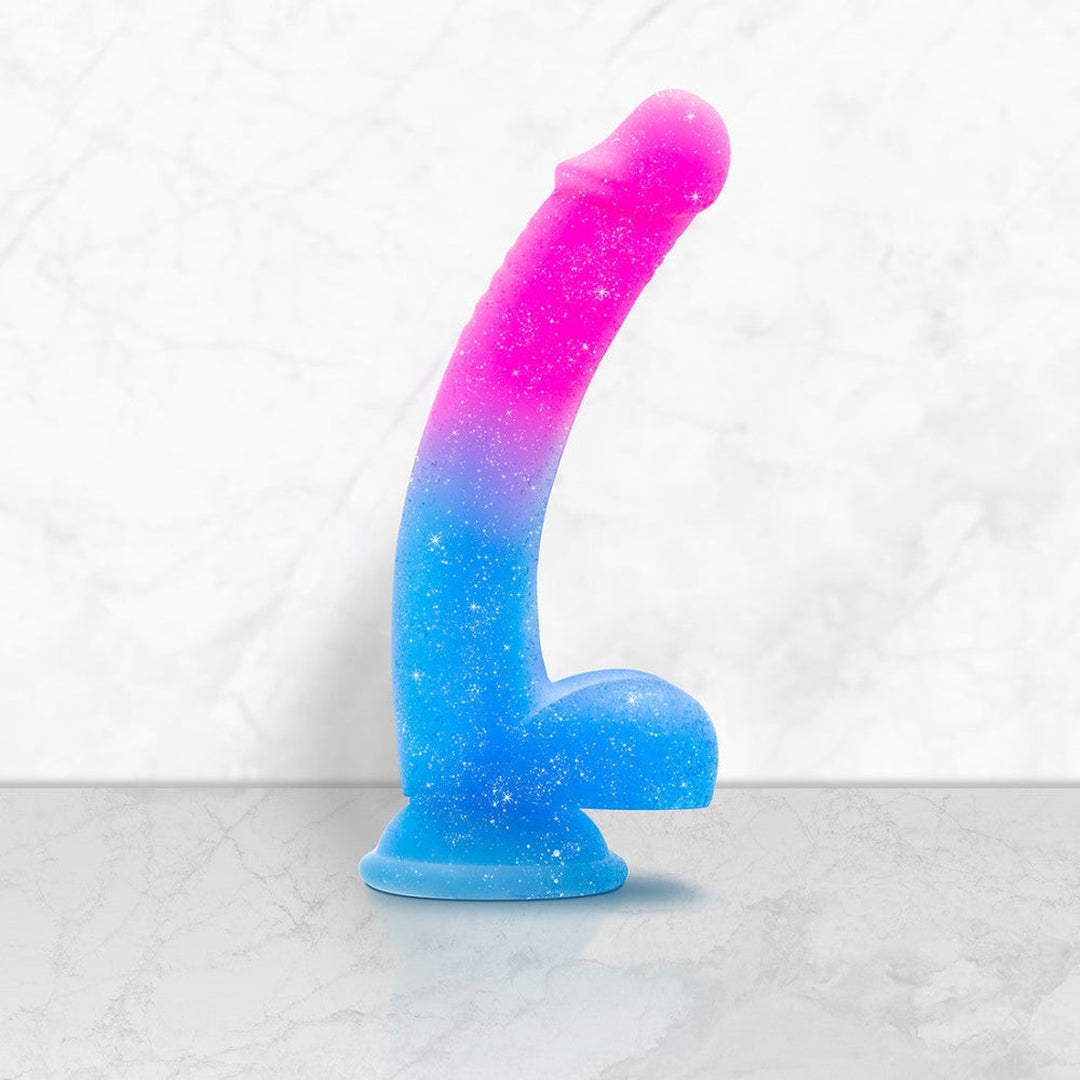 The Avant Chasing Sunsets Dildo - Mermaid by Avant stands upright on a marble surface, showcasing its colorful gradient from pink at the top to blue at the base. Crafted with Puria platinum-cured silicone, this glittery textured toy features a realistic shape and a suction cup base for added versatility.